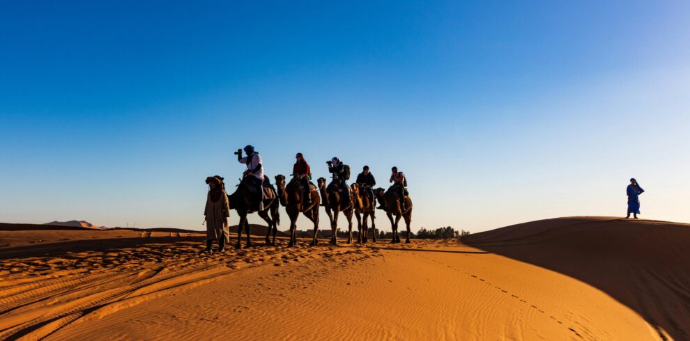 A group of travelers on camels crossing the sandy dunes of the Sahara Desert under a clear blue sky.