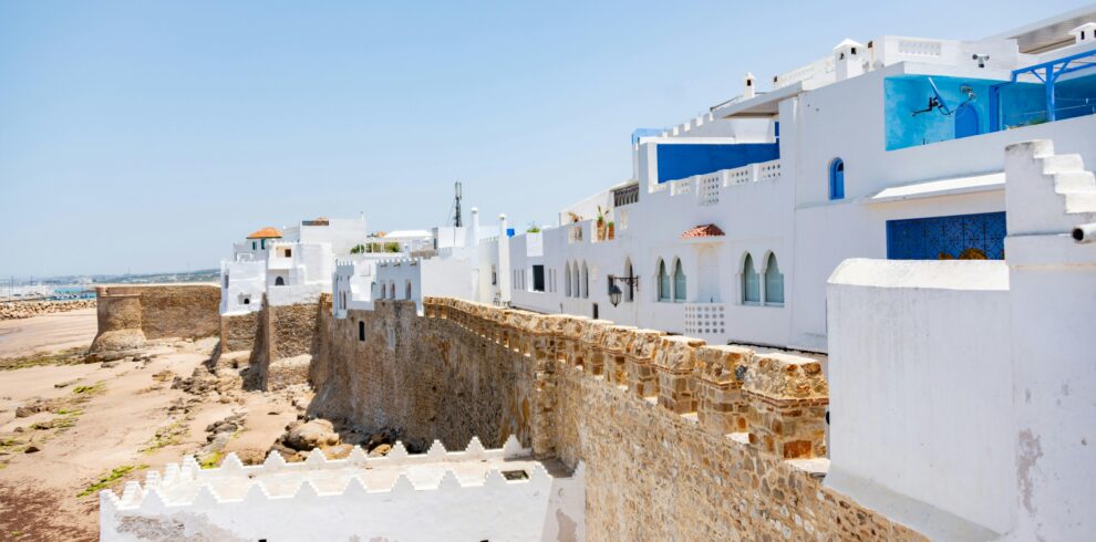 A view of a whitewashed medina with an ancient seaside fortress under a clear blue sky in Morocco.